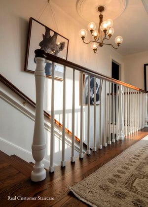 A classic indoor staircase with white balusters, a dark wood handrail, hardwood flooring, and a patterned area rug. A brass chandelier hangs from the ceiling, and framed wall art is displayed along the wall.
