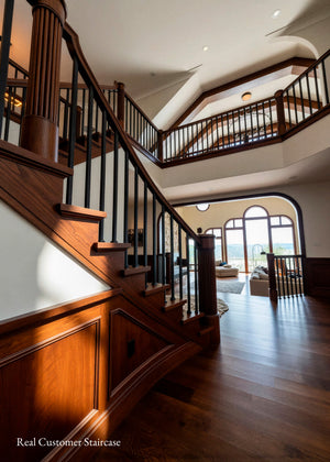 A grand wooden staircase with dark metal balusters and warm medium-brown hardwood steps, set in a spacious home interior with hardwood floors and arched windows in the background.