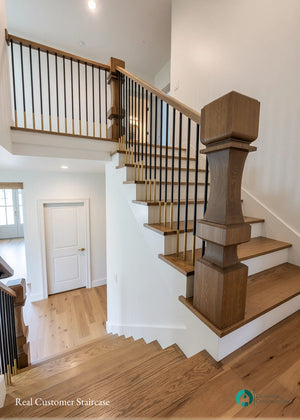 A modern wooden staircase with medium brown wood steps and handrail, paired with black metal round balusters and a substantial square wooden newel post, set against white walls in a bright home interior.