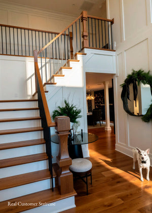 A real customer staircase featuring warm medium-brown wood steps and handrail with black metal balusters, set in a bright home interior with hardwood floors, a small glass table, and a dog on the floor.