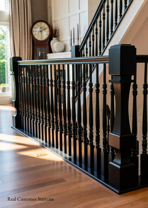A close-up view of a black wooden staircase railing with turned spindles and substantial newel posts, installed on a hardwood floor beside a staircase in a home interior.
