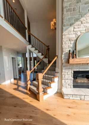A modern two-story entryway staircase with light wood treads, warm wood railings, and black metal balusters, set in a bright home with a stone fireplace and wood floors.


