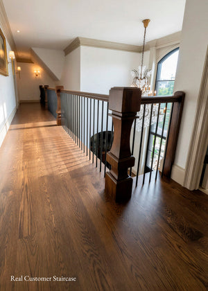 A real customer staircase shown in a home interior, featuring dark hardwood flooring, a dark wood banister with turned newel posts, white walls, and a chandelier.

