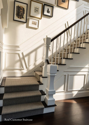 A real customer staircase with gray carpeted steps, white balusters, a dark wood handrail, white wainscoting, and framed wall art on a white wall.