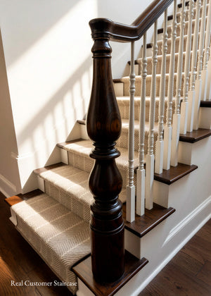 A close-up view of an indoor staircase with dark brown wooden steps and a matching dark wooden handrail, carpeted with beige woven stair runner, and supported by white balusters against white walls.