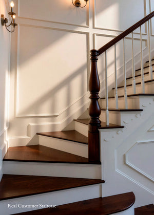 A close-up view of an indoor staircase with dark wood treads and a matching dark wood banister, set against white paneled walls with warm wall sconces casting shadows.