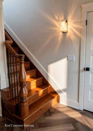 A warm-toned wooden staircase with a turned spindle banister and black metal railings, set against a white wall with a glowing wall sconce. The scene includes a white door on the right and a wood herringbone floor, with the text 'Real Customer Staircase' in the bottom-left corner.