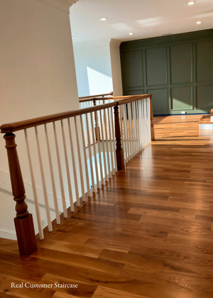 A real staircase interior with a wooden handrail and white spindles, hardwood flooring, and a dark paneled wall in the background.