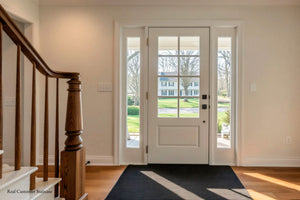 A white front entry door with two tall glass sidelight panels, shown in a bright home interior with hardwood floors, a dark runner rug, and a wooden staircase on the left. A house and trees are visible outside.