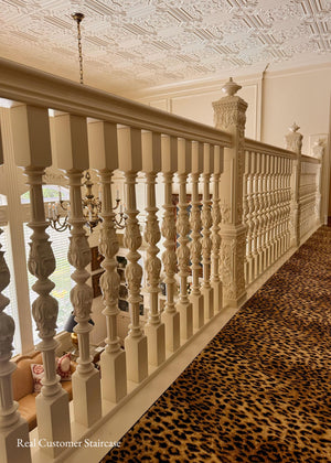 An elegant staircase railing with ornate cream-colored turned spindles and a matching handrail, installed in a home hallway with a leopard-print carpet and a decorative ceiling.
